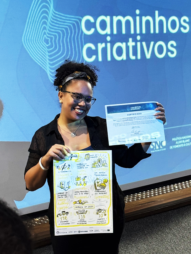 Empreendedora sorridente exibindo o certificado de participação do evento Caminhos Criativos e a prancha de facilitação gráfica do projeto Contos de Guia.
