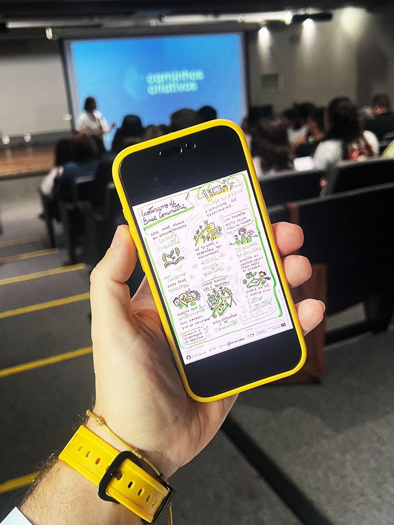 iPhone na mão de um espetador no auditório exibindo no ecrã a arte digital da facilitação gráfica sobre Ecoturismo de Base Comunitária.  