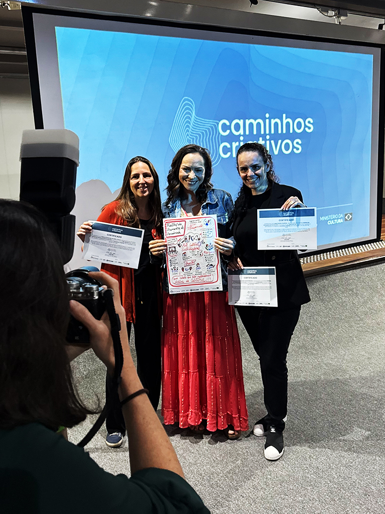 Três mulheres a posar no palco a segurar a prancha visual e certificados enquanto são fotografadas.  