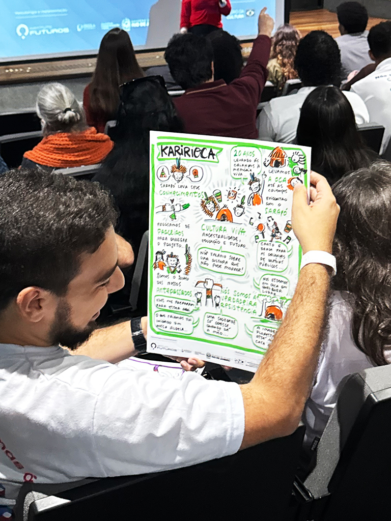 Participante na plateia a segurar e a ler a prancha de facilitação gráfica verde e ilustrada da iniciativa KaririOca.